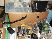 Wide view showing divided storage box with earbuds, Midland CB transceiver on cardboard box, and assorted cameras and products spread on surface.