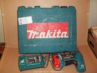 Makita cordless drill in blue-green with black handle, Makita hard carrying case, battery booster, and charger laid out on a plain surface
