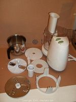 Food processor base unit with attached clear plastic bowl and feeding chute, control buttons on the side, and multiple accessory cutting blades and discs laid out beside it