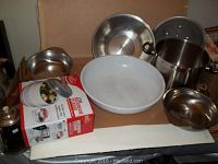 Photo of entire lot showing Handy Gourmet microwave steamer in box, stainless steel bowls, large white salad bowl, heavy bottom cook pot, and tin colander.