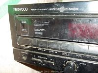 Close-up photo of the Kenwood AM-FM stereo receiver front panel showing the brand, model, input selector buttons, and speaker/output controls.