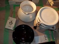 White ceramic coffee pot without lid alongside white metal lidded canister with sealing clamp, black ashtray, deck of cards pack, and number plate