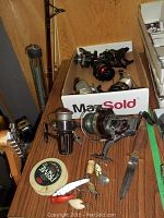 Multiple fishing reels and fishing lures displayed on wooden surface along with a fixed blade knife and fishing line spool.