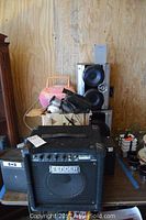 Fender Rumble 15 bass amplifier and surrounding electronic items including speakers and cameras stacked on a wooden table against a plywood wall.