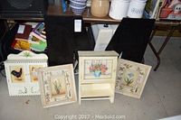 Two framed flower pictures and two painted kitchen wall cabinets with floral and rooster motifs placed on floor along with large black wooden letter M behind them.