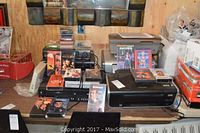 Image showing multiple printers, DVD player, CD players, VHS player and stacks of VHS and CD media on a table.