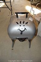 Front view of the silver Happy Griller charcoal grill with a smiling face design on the lid, showing black handle on top and legs supporting the unit.