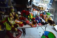 Photo showing various plush toys including a Teenage Mutant Ninja Turtle, red dinosaur, smaller stuffed animals, and toy vehicles on a white folding table.