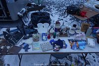 Overall view of all items on a table covered with thin snow, showing rollerblades, ice skates, ceramic mugs, bowls, novelty items, snack packs, and small household goods.