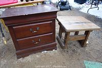 Two wooden tables outdoors: dark-finished end table with two drawers and small solid wood natural finish table side by side on ground with snow in background.