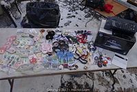 Wide overhead photo showing a large array of costume jewelry including colorful necklaces, bracelets, earrings, and angel pins spread across a white folding table outdoors on snow.