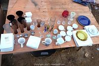 Wide view of square white ceramic plates, soup bowls, ceramic cups, mugs, various glassware including clear, colored, stemware and beer mugs arranged on a wooden surface.