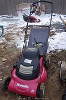 Front view of the red Homelite lawn mower with attached cloth grass catcher and foldable handle on a dirt and snow background.