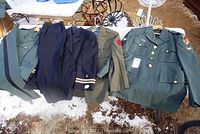 Four vintage military army uniforms displayed on hangers outdoors including two sets with pants, showing various patches and insignia.