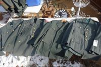 Four green vintage US Army jackets laid out on a snowy surface showing multiple gold-tone buttons and two chest pockets each.
