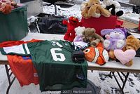 Photo showing green NY Jets jersey with #6 and Sanchez on back, assorted stuffed animals including Elmo and fish plush, and a black Polaroid camera on a folding table outside on snow.