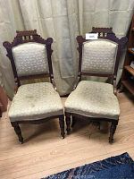 Two Victorian Eastlake side chairs with carved wooden frames and pale floral upholstery, seated side by side on wooden floor in front of curtain.