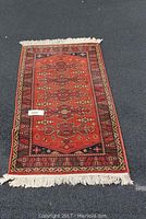 Full view of rectangular Capel Original Kashmir wool rug showing red background with detailed floral and geometric pattern, fringed ends, laid flat on asphalt.