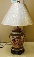 Full view of the lamp assembled, showing the white pleated plastic lampshade, oriental style ceramic ginger jar base in red and white, placed on a dark wooden stand.