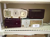 Front view of four vintage radios on a shelf, showing white Zenith radio with clock, dark brown CBS Bakelite radio, beige General Electric radio, and a small white radio in front.