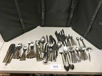 Overview photo showing a large assortment of mixed stainless flatware including forks, knives, spoons, and various serving utensils laid out on a table with black carrying cases in the background.