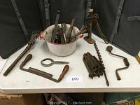 Overall view of the lot on folding table: enamel bucket with tools, screw jack, brace, auger bits, drawknife, spanner, clamp