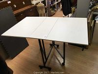 Top view of white rolling drop leaf work table fully open, supported by black metal legs and caster wheels.