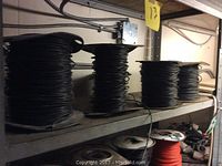 Four spools of black insulated wire on metal shelf, varying sizes, two labels visible on some spools