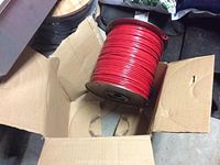 Spool of red wire in open cardboard box on cement floor with some packing material visible.