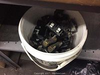 White plastic bucket filled with multiple black fuse blocks containing glass fuses, stored on a shelf in a warehouse or storage area.