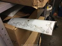 Photo showing a white curved bathroom light shade with silver decorative scrollwork design partially protruding from an open box on a shelf.