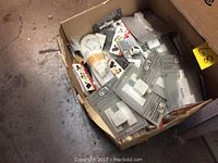 Cardboard box full of electrical items including packaged switches and sockets along with other loose electrical components on a concrete floor.