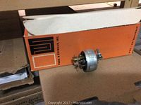 Close up of a single metallic electrical ground plug in front of an orange box from M & J Seymour Inc.