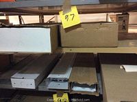 Fluorescent light fixtures placed on metal shelving, some partially out of boxes, showing rectangular white metal housing.