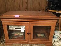 Front view of the wooden TV stand showing double glass doors and wooden frame, displaying some electronic devices inside through glass.