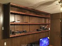View of the wooden wall-mounted three-tier shelf, showing the scalloped edging and items on the middle and lower shelves against a wood-paneled wall.
