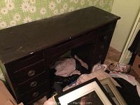Front view of dark wooden sewing table with nine drawers and visible wear, scratches on surface, and fabric items on the floor nearby.