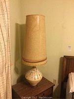 Full view of ceramic table lamp with mosaic tiles on base and textured beige lampshade placed on dark wood rectangular end table next to bed.