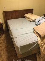 Full view of full bed with wooden headboard, fitted blue sheet on mattress, pillows and blanket on bed