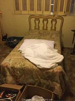 Full view of the twin bed with floral bedspread, cream colored decorative wooden headboard, and part of surrounding room visible including carpet and wall.