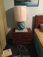 Wooden telephone table placed beside a bed, topped with a blue elephant head lamp base (lamp not included). The table displays surface wear and a lace doily under the lamp.