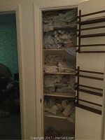 Photo of wooden built-in closet shelves filled with assorted off-white and muted color linens folded and stacked, viewed with closet door partially open.