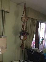 Two hanging ceramic pots with faux flowers and macrame braided supports shown indoors near window and curtains.
