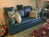 Front view of dark green/teal two seater sleeper couch with five pillows on a green carpeted floor.