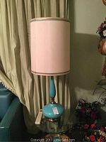 Pair of matching turquoise vintage lamps on a glass table, tall beige shades with aqua trim, visible curtain backdrop and floral decoration nearby