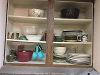 Cabinet showing assorted ceramic and glass dishes including bowls, plates, mugs, and blue plastic measuring cups.