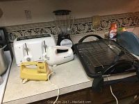 Photo showing the two hand mixers (white and yellow), white toaster, black electric grill on counter with wall outlet and decorative wallpaper border