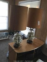 Pair of vintage table lamps on a wooden surface near windows with beige lampshades and black and white ceramic bases featuring scrollwork patterns.