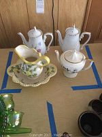 Top down view of three white teapots with floral and gold detail, along with a ceramic water bowl with underplate.