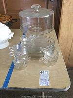 Full view of the glass cake plate with dome lid, two glass cups with handles, two glass saucers, and a plate on a table.